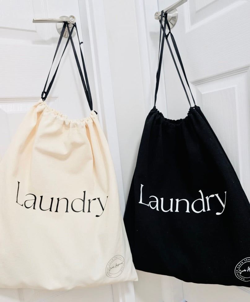Two drawstring bags labeled 'Laundry' hanging on a rack. One black and one cream / beige