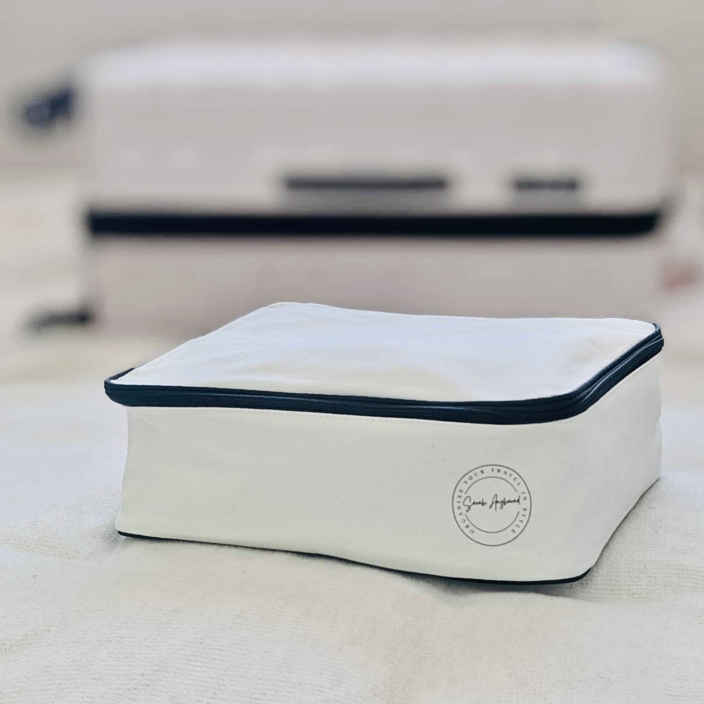 Cream travel packing cube with black trim on a light surface, with a suitcase in the background.