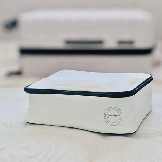 Cream travel packing cube with black trim on a light surface, with a suitcase in the background.