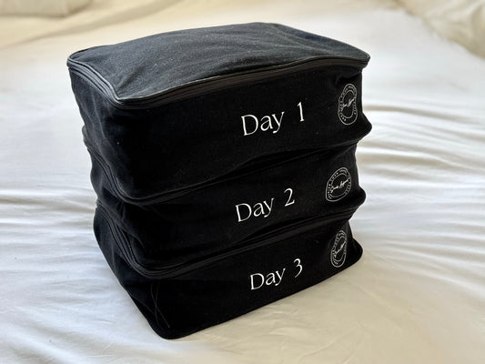 Stack of black travel accessories labeled 'Day 1', 'Day 2', and 'Day 3' on a white bed.