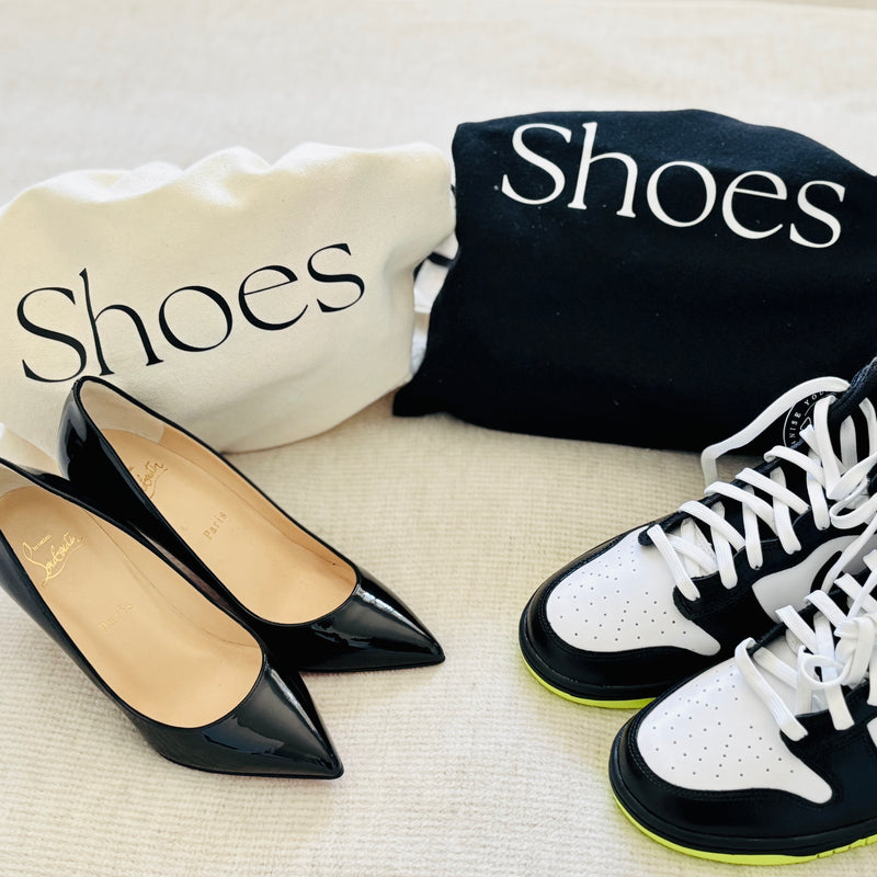 Black pointed-toe shoes and white sneakers with black soles on a light surface, with 'Shoes' bags in the background.